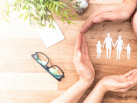 Image of a pair of glasses on a table and four hands forming a circle around a paper cutout of a family of four.
